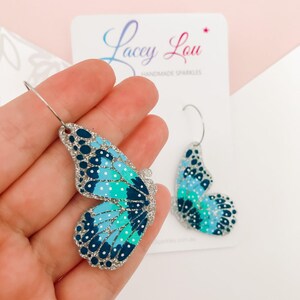 May include: Butterfly wing earrings in shades of blue and teal with white polka dots and silver glitter accents. The earrings are on a white card with the text "Lacey Lou Handmade Sparkles".