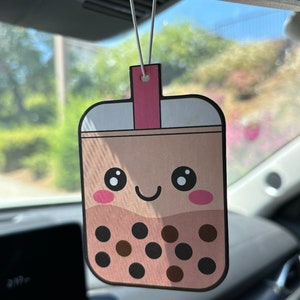 May include: A brown and white car air freshener shaped like a boba tea cup with a pink lid and a smiling face. The cup is filled with brown boba pearls.