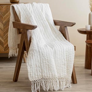 May include: A white cotton throw blanket with a crocheted fringe, draped over a wooden chair. The blanket has a subtle textured pattern and is ideal for adding a touch of warmth and style to any room.