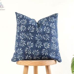 Op de afbeelding: Een vierkant, marineblauw sierkussen met een wit bladerpatroon. Het kussen ligt op een kleine, ronde houten kruk. Een decoratief woonaccessoire.