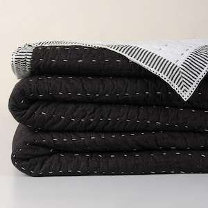 Könnte beinhalten: Ein Stapel aus drei schwarzen Quilts mit weißen Nähten. Der obere Quilt hat einen weiß-schwarz gestreiften Rand. Die Quilts sind ordentlich gefaltet und zeigen die Textur und das Design. Die Quilts sind wahrscheinlich für ein Bett oder Sofa.