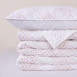 Peut inclure: Une pile de literie blanche, comprenant un oreiller et des couettes pliées, avec des coutures rouges. L'oreiller est en haut, suivi des couettes pliées. Les couettes et l'oreiller ont un design simple mais élégant.
