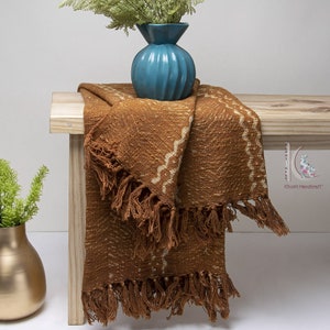 May include: A brown throw blanket with a white geometric pattern and fringe. The blanket is draped over a wooden bench with a blue vase of greenery on top.