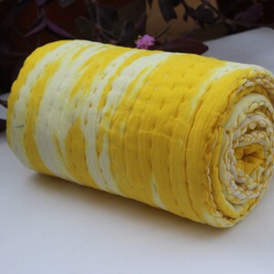 Op de afbeelding: Een gele en witte kantha-deken opgerold. De deken heeft een tie-dye-patroon en is gemaakt van katoen.