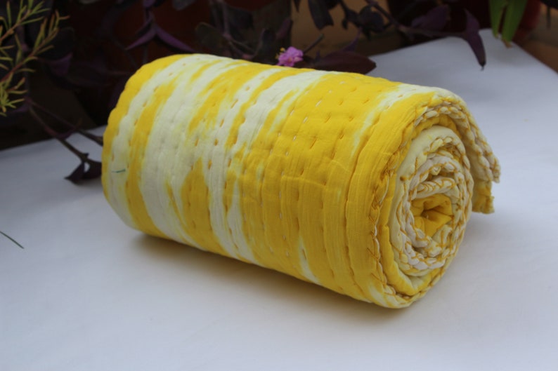Op de afbeelding: Een gele en witte kantha-deken opgerold. De deken heeft een tie-dye-patroon en is gemaakt van katoen.