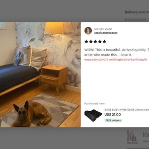 May include: A black and white kantha quilt with a natural design is displayed on a bed in a bedroom. The quilt is on a wooden bed frame with a brown dog lying on a gray and white patterned rug in front of the bed. The bedroom has a white wall with a floral wallpaper accent wall. A small wooden nightstand with a lamp is next to the bed.