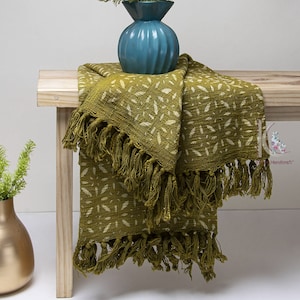 May include: A green throw blanket with a white leaf pattern and fringe. The blanket is folded and draped over a wooden bench.