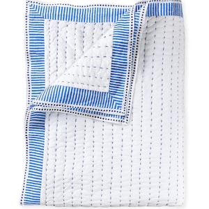 May include: A white quilt with blue striped borders and vertical blue stitching. The quilt has a folded corner, revealing the interior stitching pattern. The border features a series of blue stripes and small blue dots. This bedding item is suitable for a bed or couch.