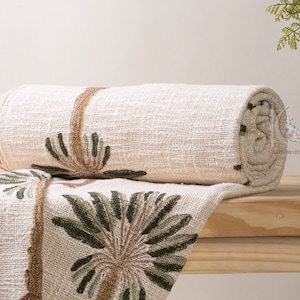 以下が含まれることがあります： 緑と茶色の刺繍で飾られた白い毛布。毛布は丸められて木製の表面に置かれています。