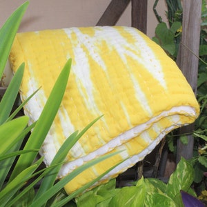 Op de afbeelding: Een gele en witte tie-dye kantha quilt gevouwen in de helft. De quilt is gemaakt van katoen en heeft een zacht, getextureerd oppervlak.