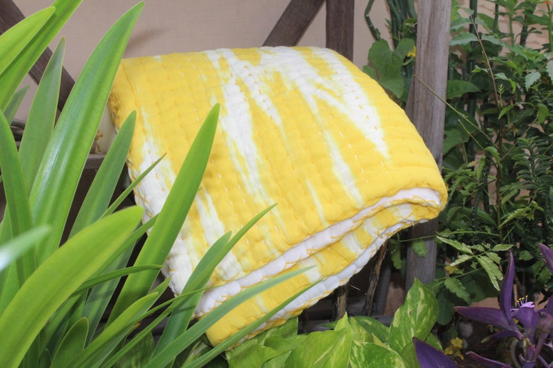 Op de afbeelding: Een gele en witte tie-dye kantha quilt gevouwen in de helft. De quilt is gemaakt van katoen en heeft een zacht, getextureerd oppervlak.