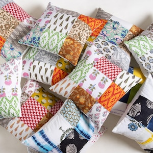 May include: A collection of square throw pillows with patchwork designs. The pillows feature a variety of patterns, including floral, geometric, and abstract prints in colors like red, orange, green, and blue. The pillows are arranged in a pile.