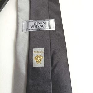 Puede incluir: Corbata de seda negra con una etiqueta plateada que dice "Gianni Versace" y una etiqueta plateada más pequeña con el logotipo de Versace, una cabeza de Medusa dorada.