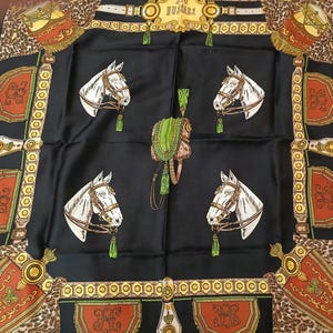 Puede incluir: Un pañuelo cuadrado negro con cuatro cabezas de caballo blancas con bridas marrones y borlas verdes. El diseño incluye una silla de montar verde central, esquinas con estampado de leopardo y bordes decorativos con patrones geométricos rojos y dorados y el texto LES HUSSARDS.