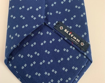 May include: A blue necktie with a repeating pattern of small, light blue squares. The tie has a textured surface and a label with text and floral designs. The tie is made of a smooth fabric.
