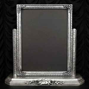 May include: A silver-toned, rectangular picture frame with ornate floral and scrollwork detailing. The frame has a black backing and is supported by a base with a floral design. The frame is designed to stand upright.