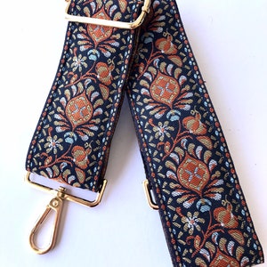 May include: An adjustable strap with a black and gold patterned design and gold metal hardware. The strap is made of woven fabric and features a floral pattern.