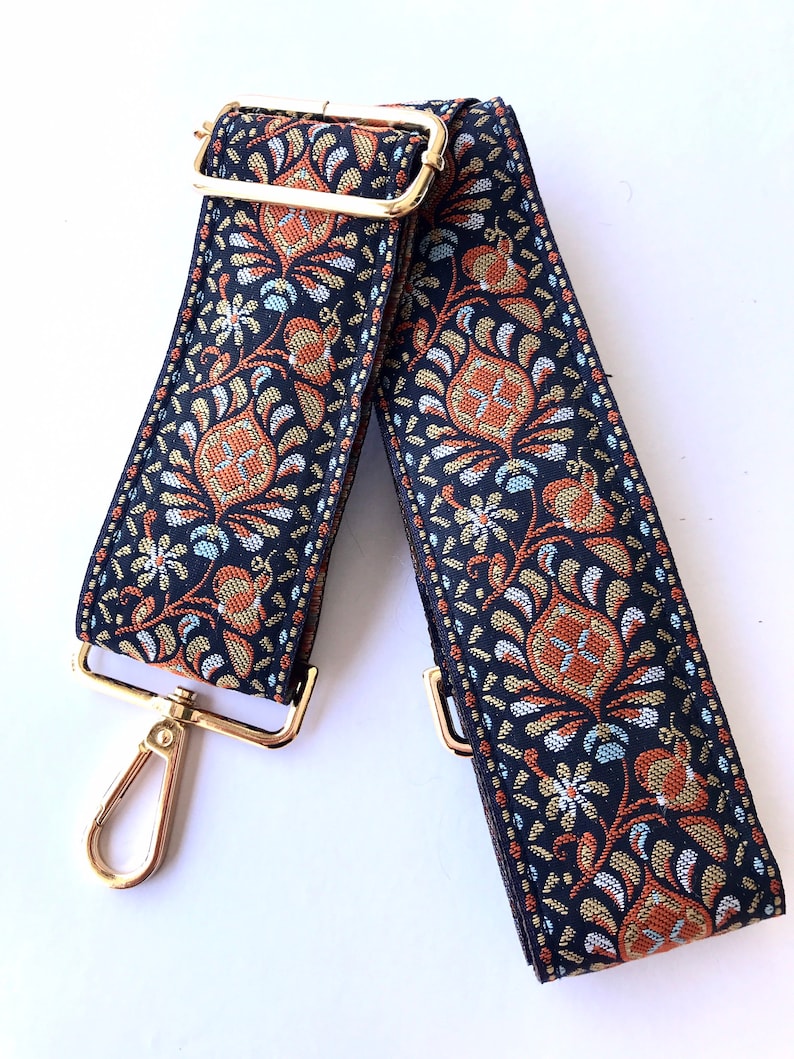 May include: An adjustable strap with a black and gold patterned design and gold metal hardware. The strap is made of woven fabric and features a floral pattern.