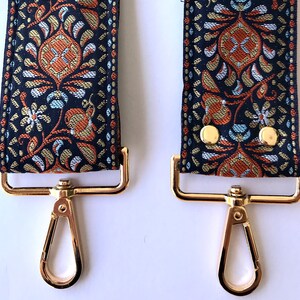 May include: Close-up of two gold-tone metal clasps with a black and gold patterned fabric strap. The strap has a floral design with orange, blue, and gold accents.
