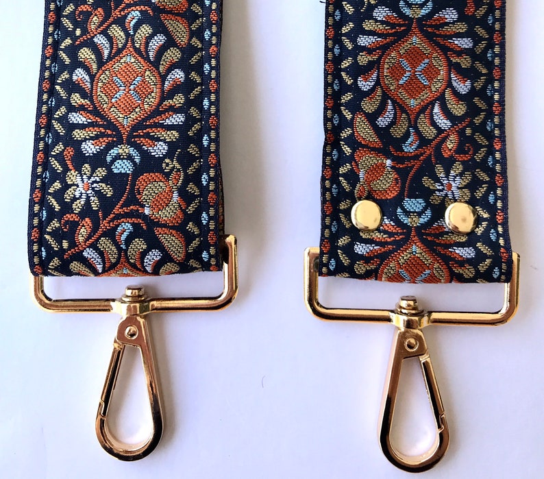 May include: Close-up of two gold-tone metal clasps with a black and gold patterned fabric strap. The strap has a floral design with orange, blue, and gold accents.