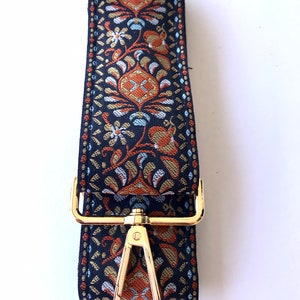 May include: A black and gold patterned fabric strap with a gold metal clasp and swivel hook. The strap is designed for a bag or purse.