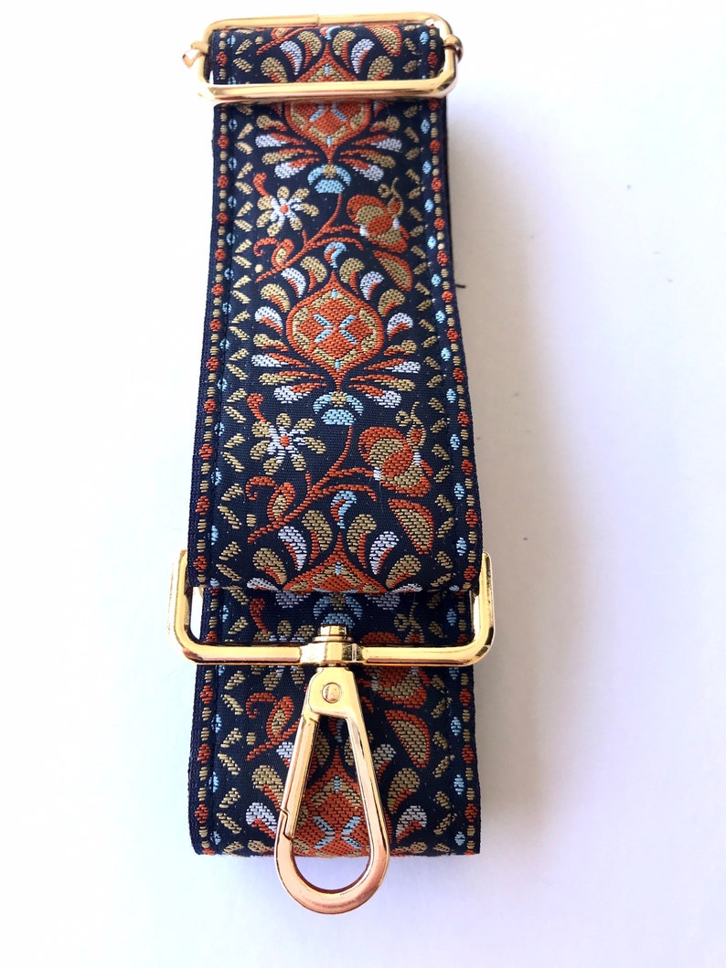 May include: A black and gold patterned fabric strap with a gold metal clasp and swivel hook. The strap is designed for a bag or purse.