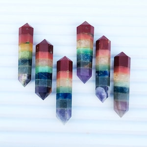 Op de afbeelding: Zes zeven chakra kristallen staven, elk met een puntige punt en gelaagd met zeven verschillende gekleurde stenen, die de zeven chakra's voorstellen. De staven zijn in een halve cirkel op een wit oppervlak gerangschikt.