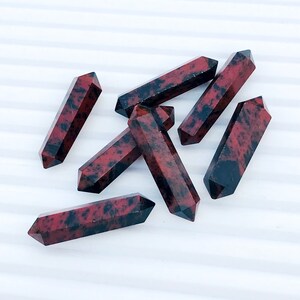 May include: Seven red and black gemstone points, also known as red jasper points, are arranged on a white surface. The points are all roughly the same size and shape, and they have a natural, unpolished finish.