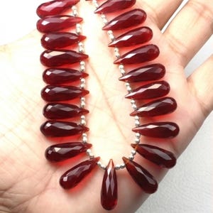 Puede incluir: Un collar con gemas facetadas en forma de lágrima, de color rojo intenso, ensartadas en una cadena de plata. Las gemas están dispuestas en un patrón simétrico, con un solo colgante de lágrima más grande.