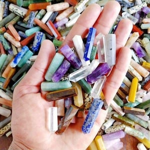 Puede incluir: Una mano sosteniendo una variedad de varitas de cristal de colores. Las varitas están hechas de diferentes tipos de piedras, incluyendo amatista, cuarzo transparente y ojo de tigre.