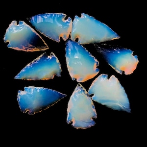 May include: A collection of nine iridescent blue and white arrowhead shaped stones. The stones have a smooth, polished surface and are arranged on a black background.
