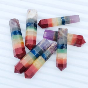 May include: Seven chakra crystal wands, each with a different color representing a chakra, arranged on a white surface.