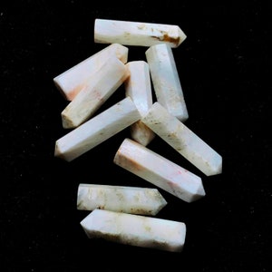 Könnte beinhalten: Eine Gruppe von acht weißen, undurchsichtigen, spitzen Kristallstäben mit einem leichten Rosaton. Die Stäbe sind in einem zufälligen Muster auf einem schwarzen Hintergrund angeordnet.