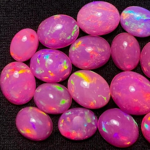 May include: A collection of pink opal cabochons with iridescent flashes of colour. The oval-shaped stones are arranged in a cluster on a black background.
