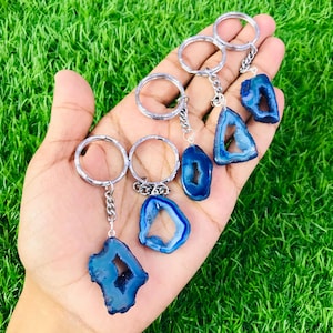 Op de afbeelding: Een verzameling blauwe agaat stenen sleutelhangers met zilverkleurige metalen ringen en kettingen. De agaatstenen hebben een natuurlijke, onregelmatige vorm met een donkerblauw centrum en een lichtere blauwe rand. Op een groene achtergrond.