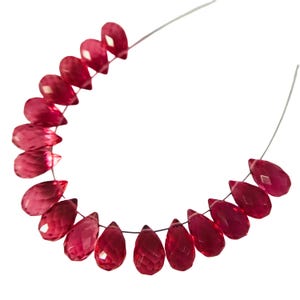 May include: Strand of faceted, teardrop-shaped beads in a deep red hue. The beads are strung on a thin, dark thread, with a few beads visible at each end. The beads have a glossy finish and are evenly spaced.