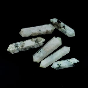 May include: Six polished, double-terminated moonstone wands. The wands are primarily white with gray and black inclusions. The wands are arranged on a black background. Each wand has a hexagonal shape.