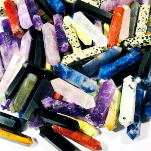 Può includere: Una collezione di bacchette di cristallo appuntite, lucidate e colorate, in vari colori, tra cui viola, blu, verde, rosso, nero e bianco. Le bacchette sono disposte in modo casuale su una superficie bianca.