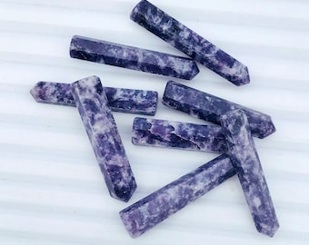 Könnte beinhalten: Sechs violette und weiße Amethyst-Stabformen, auch als Spitzen bekannt, liegen auf einer weißen Oberfläche verstreut. Die Stäbe sind alle ungefähr gleich groß und geformt, mit einer spitzen Spitze und einer abgerundeten Basis.