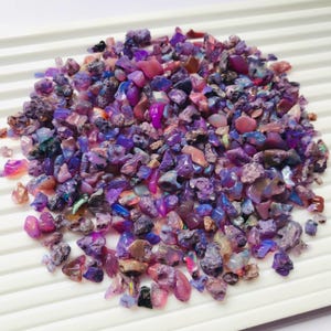 May include: A pile of raw, uncut opal gemstones in shades of purple, pink, and blue. The opals are of various sizes and shapes, with a rough, natural texture. The gemstones are displayed on a white, ridged surface.