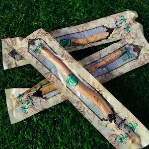 Op de afbeelding: Verschillende verpakte miswak-stokken zijn gerangschikt op een groen oppervlak. Elke stok is verzegeld in een doorzichtige plastic verpakking met een decoratief beige en bruin patroon. De verpakking bevat groene labels en Arabische tekst.