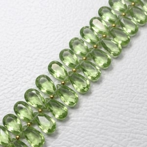 May include: Close-up of a strand of light green, teardrop-shaped gemstones. The gems are strung together with small gold beads, creating a delicate and elegant jewelry piece. The background is a soft, white surface.
