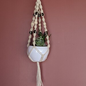 May include: A white ceramic planter with a green succulent plant hanging from a macrame plant hanger with black and white wooden beads.