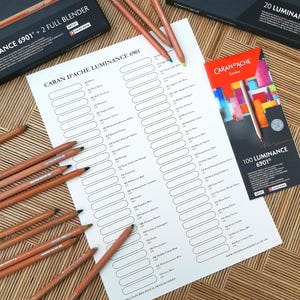May include: A white sheet of paper with a color chart for Caran d'Ache Luminance 6901 colored pencils. The chart has 100 numbered circles for each color. The paper is surrounded by wooden colored pencils and boxes of Caran d'Ache Luminance pencils.
