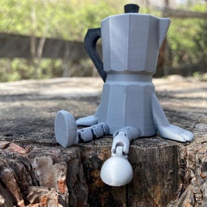 May include: A grey ceramic coffee pot with a black handle and spout, shaped like a robot with arms and legs. The robot is sitting on a tree stump.