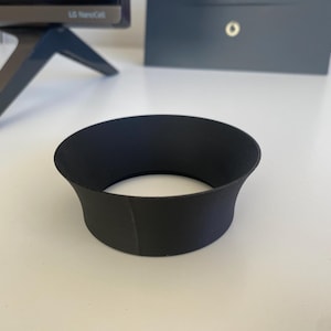 Könnte beinhalten: Ein schwarzes, zylindrisches Objekt mit einem ausgestellten oberen Teil und einer breiten Öffnung. Das Objekt besteht aus einem matten Material und liegt auf einer weißen Oberfläche. Die Form deutet darauf hin, dass es sich um eine Sonnenblende oder ein ähnliches Zubehör handeln könnte.