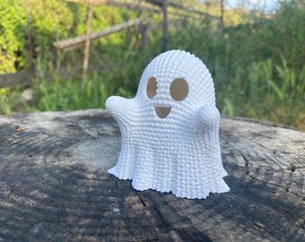May include: A white ghost figurine with a textured surface, cut-out eyes and a smiling mouth. The Halloween decoration is on a weathered wooden surface, with a blurred green background.