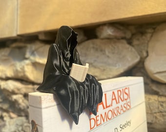 May include: A black 3D-printed figurine of a cloaked figure reading a book. The figure is perched on a book with red and white text. The background features a stone wall and a wooden ledge.