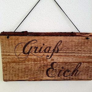 Könnte beinhalten: Ein rustikales Holzschild mit dem Text "Griass Eich", der in die Oberfläche gebrannt ist. Das Schild hängt an einer schwarzen Schnur.