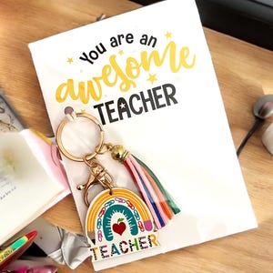 May include: A white card with the text "You are an awesome TEACHER" and a rainbow keychain. The keychain has a rainbow design with a heart and the word "TEACHER". A colorful tassel hangs from the keychain.
